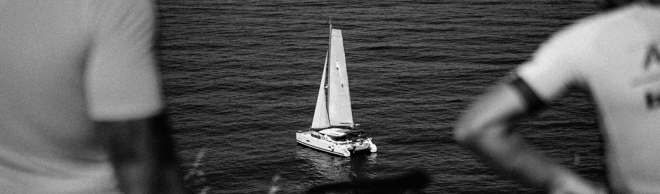 A catamaran under sail, seen past two people watching from the shore