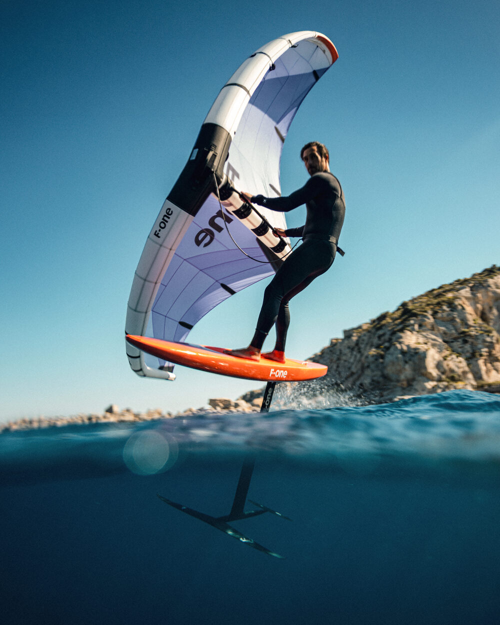 Wing foiler lifting off the water beside a rocky headland