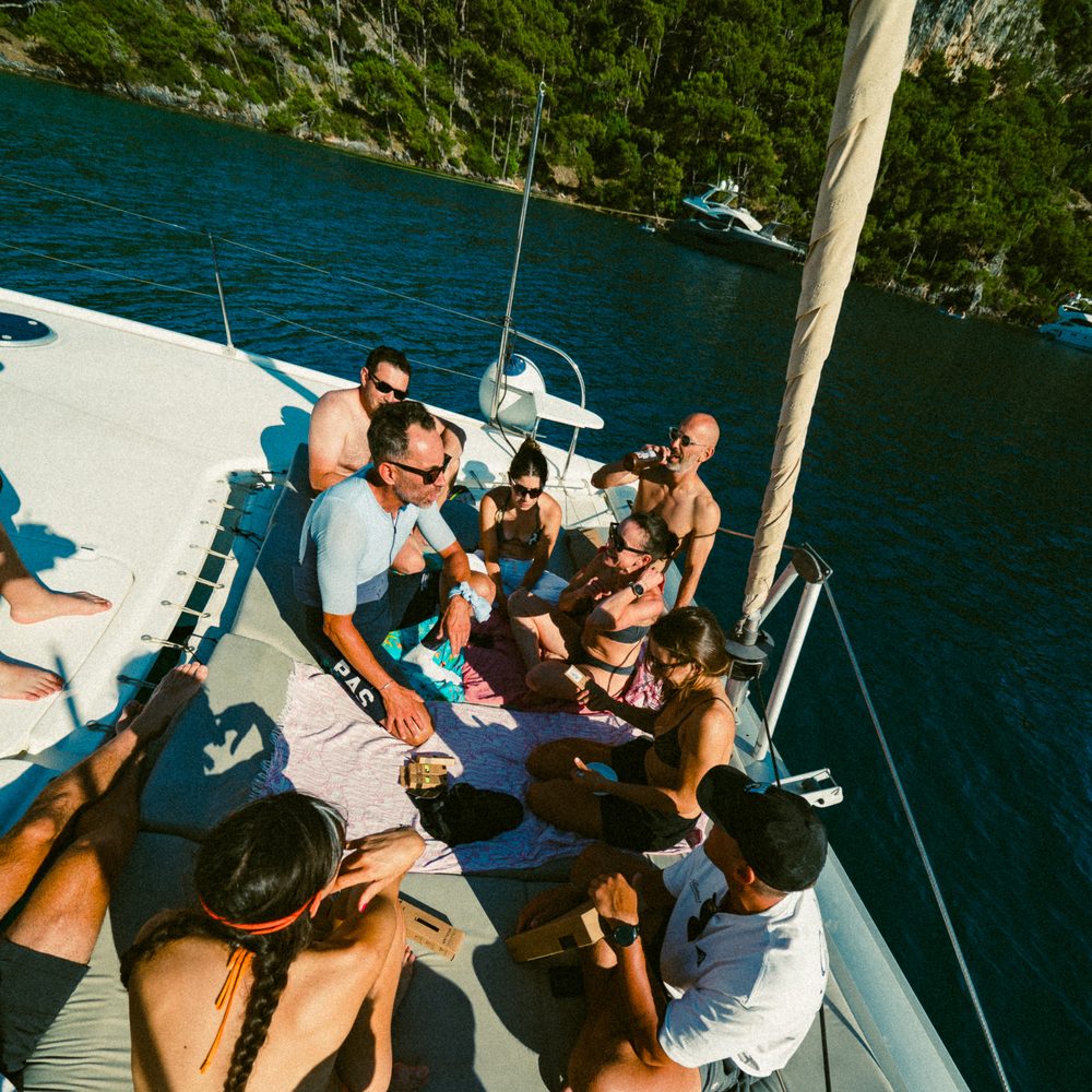 Riders resting on the foredeck of the catamaran in a pine-lined bay