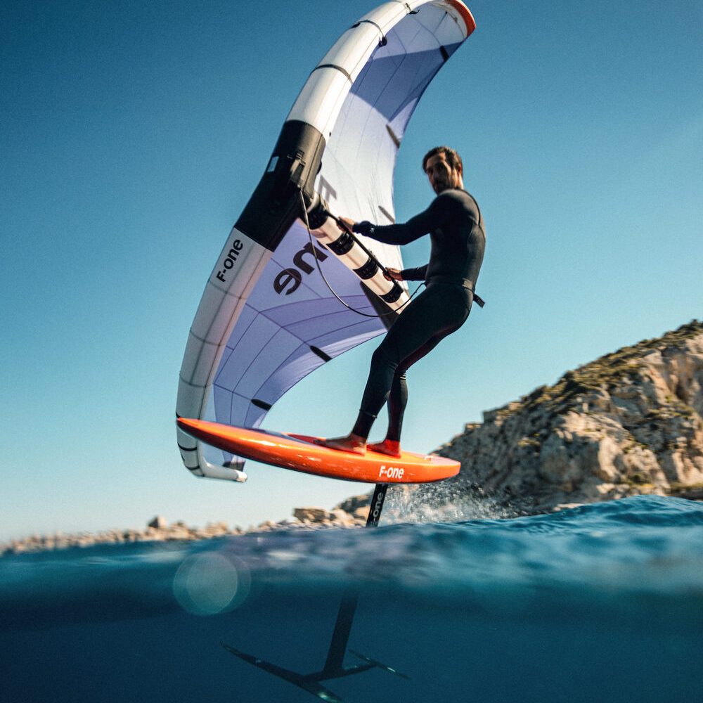 A wing foil rider lifting clear of the water beside a rocky shore
